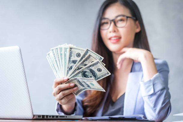 Beautiful business woman sitting at home office desk  using a laptop and smiling while working with banknote hold in hand, and and be attractive cheerful model using computer. Indoors - Photo, Image