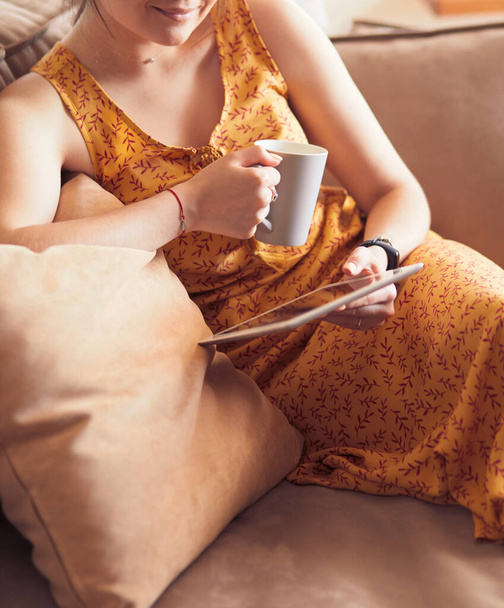 Young woman uses tablet while relaxing at home with coffee on the sofa couch - Foto, Imagen