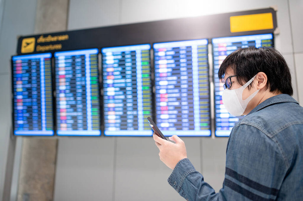 Asijské muž turista nosí masku kontrolu letu od příletu odletu palubě pomocí smartphone na letišti terminálu. Koronavirus (COVID-19) pandemická prevence při cestování. Koncept sociálního distancování - Fotografie, Obrázek