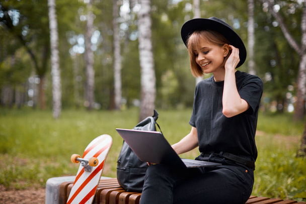 Creativo ridere felice ragazza blogger lavoro sul computer portatile sui contenuti seduti sulla panchina con skateboard e zaino nel parco - Foto, immagini