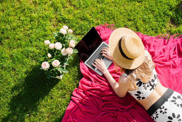 A young girl blogger in a hat working on a laptop with empty screen in the summer on the lawn, photo for a blog or ad - Foto, Imagen