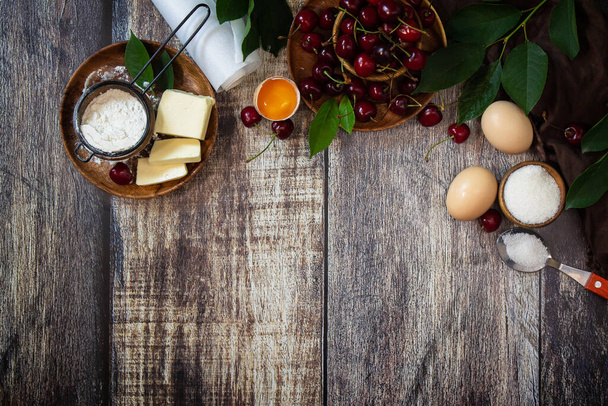Εποχιακό ψήσιμο. Υλικά για κερασόπιτα (κόκκινα κεράσια, αλεύρι, αυγά, ζάχαρη και βούτυρο) σε ένα ρουστίκ ξύλινο τραπέζι. Κάτοψη επίπεδου δαπέδου. Αντιγραφή χώρου. - Φωτογραφία, εικόνα