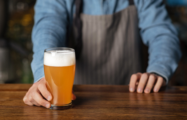 Una pinta de cerveza artesanal. Barman en delantal y camisa azul poner en vidrio de mesa de madera de ale
 - Foto, imagen