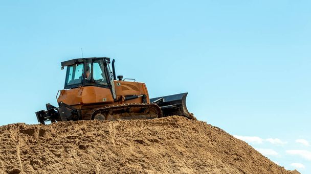 gele bulldozer op een grote stapel zand tegen een blauwe lucht - Foto, afbeelding