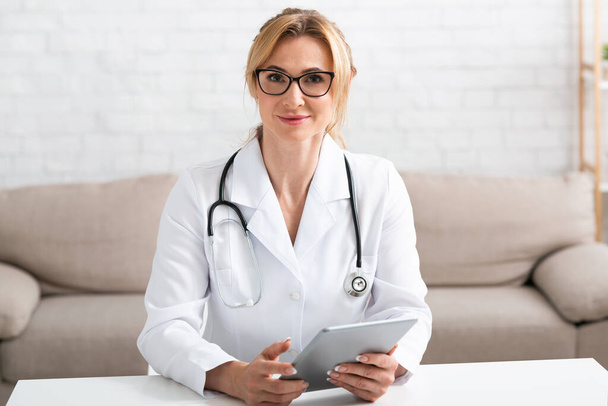 Unterstützung aus der Heimat. Ärztin im weißen Kittel mit Tablette in der Hand sitzt am Tisch - Foto, Bild
