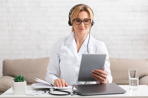 Online-Konferenz und Multitasking. Ärztin mit Kopfhörer blickt auf Tablet - Foto, Bild