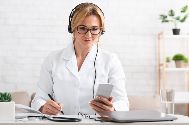 Writes patients to visit. Woman in white coat with headphones, smartphone and laptop makes notes in notebook - Fotografie, Obrázek
