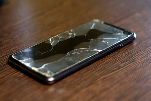 Black smartphone lies on a wooden table with a broken screen. - Photo, image