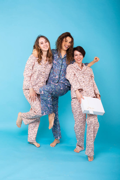 Portrait of three beautiful young girls wearing colorful pyjamas having fun during sleepover isolated over blue background. Pajama party and hen-party concept - Photo, Image