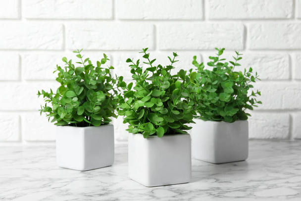 Beautiful artificial plants in flower pots on white marble table near brick wall - Фото, изображение