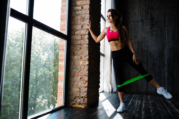 Full-length portrait of a sportswoman who put on her calves a green elastic tape to strengthen her leg muscles against a panoramic window - Photo, Image