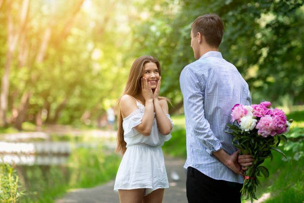 Affectionate boyfriend hiding bouquet of flowers for his beloved girlfriend behind his back at park, empty space - Photo, Image