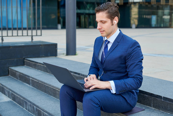 Happy successful young businessman working on modern laptop, using earphones and sitting on stairs outside skyscrapers - Foto, Imagem