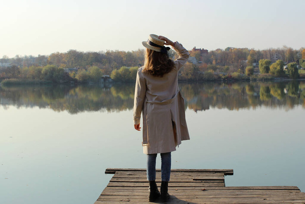 Young girl wearing a beige trench coat, jeans and black boots, standing on a wooden bridge and looking to the beautiful landscape. Young girl standing near the lake. People, nature, travel concept. - Photo, Image