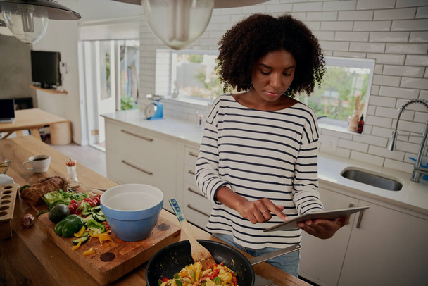 Young african woman watching recipe in digital tablet while cooking lunch in modern kitchen - Photo, Image