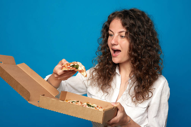 Shot of young hungry curly haired lady opens mouth widely with delicious slice of pizza, wants to eat, dressed in white shirt, models against blue studio background. Positive woman with junk food.  - Foto, imagen