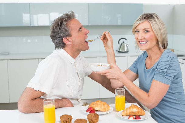 Woman feeding man at breakfast table in kitchen - Foto, afbeelding