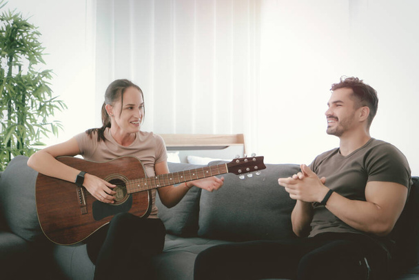 Друзі проводять час разом вдома, жінка співає пісню і грає на гітарі на дивані у вітальні, чоловік слухає музику, відпочиває і розслабляється вдома
 - Фото, зображення