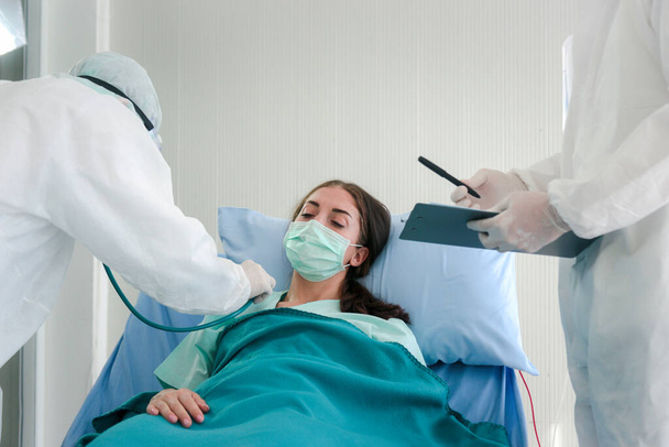 Инфицированная пациентка в маске лежит в постели в комнате для лечения заболеваний, врач в защитной одежде заботится о больных в карантине в больнице
 - Фото, изображение