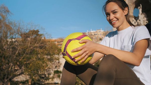 Chica deportiva bastante sonriente con el fútbol mirando alegremente en la cámara descansando en el parque después del entrenamiento
 - Foto, Imagen