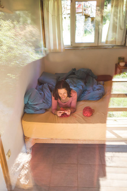 Mulher tricota deitado em uma visão de cama por um painel de janela, As mãos de mulher tricotam um chapéu, A menina está envolvida na criatividade durante a quarentena, Handmade, passatempo predileto em casa
. - Foto, Imagem