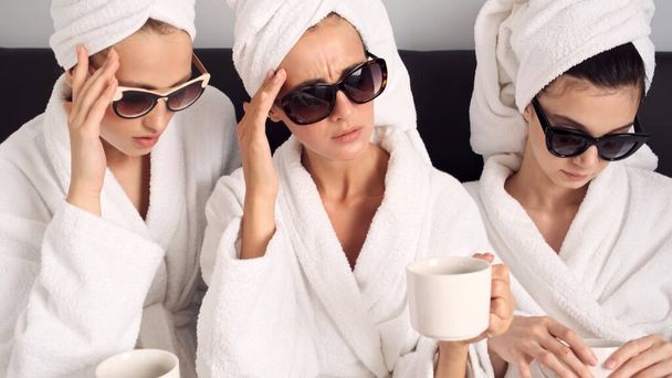 Feldúlt lányok fürdőköpenyben és napszemüvegben, kávéscsészékkel, fejfájással a buli után a hotelszobában. Szomorú kifejezés. - Fotó, kép