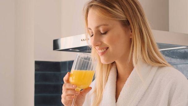 Close up pretty smiling blond girl in white bathrobe happily drinking orange juice on kitchen at home. Attractive girl having breakfast - Photo, image