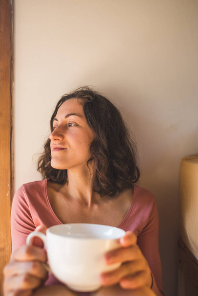 Una mujer está disfrutando del café de la mañana en casa, Retrato de una morena sonriente con una taza de bebida de la mañana, Chica está tomando té
. - Foto, imagen