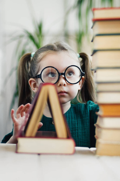 Üçgen şeklinde bir kitap diğerinin üzerinde duruyor. Arka planda yuvarlak gözlüklü küçük kız. İlköğretim - Fotoğraf, Görsel