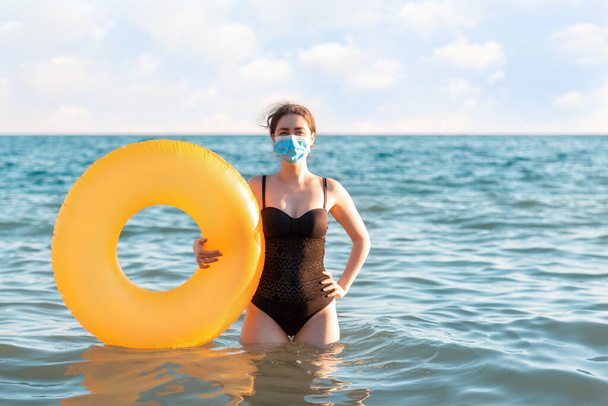 Distância social e proteção contra vírus. Uma mulher em um maiô e máscara médica posa com um círculo inflável em pé no mar. No fundo, o mar e o céu, o horizonte. Espaço de cópia
. - Foto, Imagem