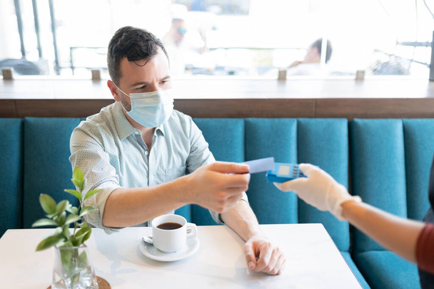 Homme adulte moyen masqué effectuant le paiement par carte de crédit au café - Photo, image