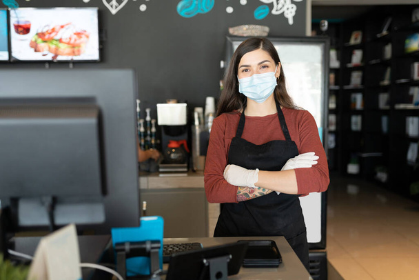 Portrét ženské baristky s obličejovou maskou při stání v kavárně se zkříženýma rukama - Fotografie, Obrázek