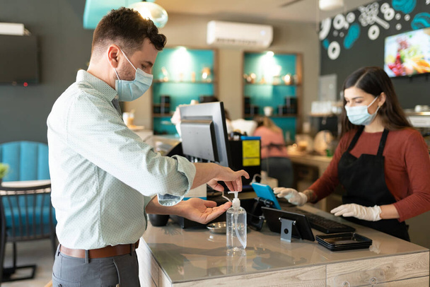 Homem caucasiano aplicando desinfetante na mão no balcão de check-out no café durante a crise do coronavírus
 - Foto, Imagem