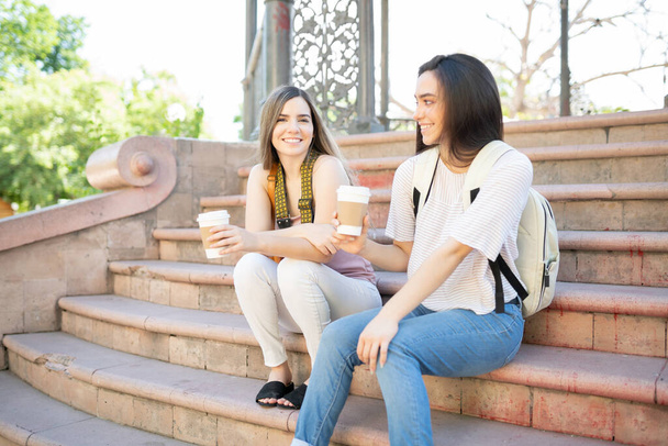Des jeunes femmes souriantes avec des tasses à café jetables assises sur les marches du parc pendant le week-end - Photo, image