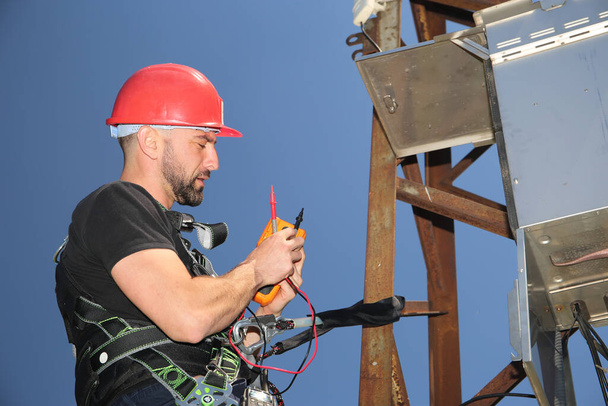 Tehnician in red helmet and safety belt with multimeter on the electric pole - Photo, Image