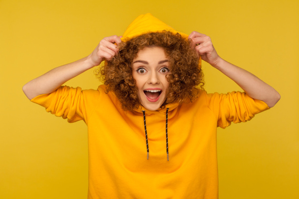 Portrait of funny amazed girl with curly hair wearing urban style hoodie, putting on hood and looking surprised at camera, youth sport style female fashion. studio shot isolated on yellow background - Fotografie, Obrázek