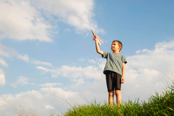 Ένα μικρό αγόρι εκτοξεύει ένα χάρτινο αεροπλάνο στον αέρα. Ένα παιδί εκτοξεύει ένα χάρτινο αεροπλάνο. Ευτυχισμένο παιδί που παίζει με χάρτινο αεροπλάνο. - Φωτογραφία, εικόνα
