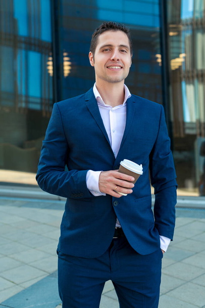 Smiling business man in suit holding cup of coffee on background with skyscrapers - Foto, Bild