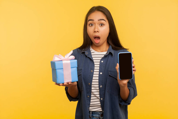 Wow, gift bonus for mobile user! Portrait of shocked girl in denim shirt holding wrapped present and cell phone with empty display, mock up for app advertise. studio shot isolated on yellow background - Zdjęcie, obraz