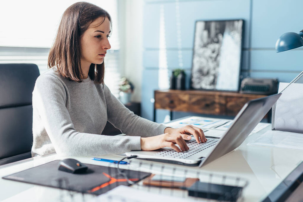 Een vrouw die een laptop gebruikt terwijl ze aan haar bureau zit. Zakenvrouw op kantoor werkt aan laptop - Foto, afbeelding