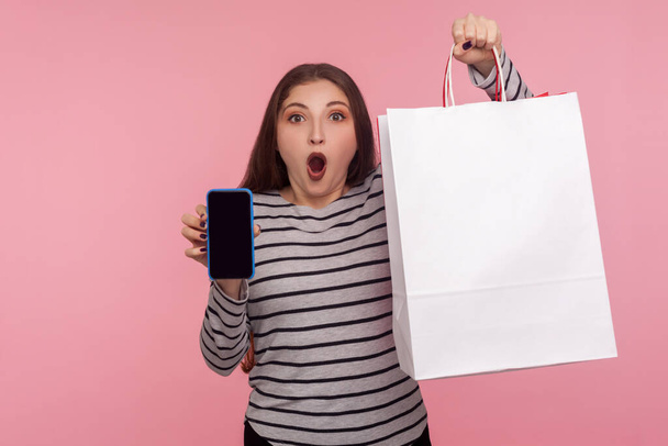 Unbelievable online store app! Portrait of shocked woman in sweatshirt holding shopping bag and cell phone, showing mobile device with blank mock up for advertise. studio shot, yellow background - Photo, image