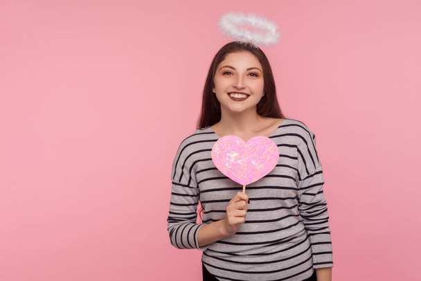 Happy Valentine's day! Angelic woman with holy nimbus holding pink paper heart and smiling to camera, demonstrating love care, romantic feelings. indoor studio shot isolated on pink background - Zdjęcie, obraz