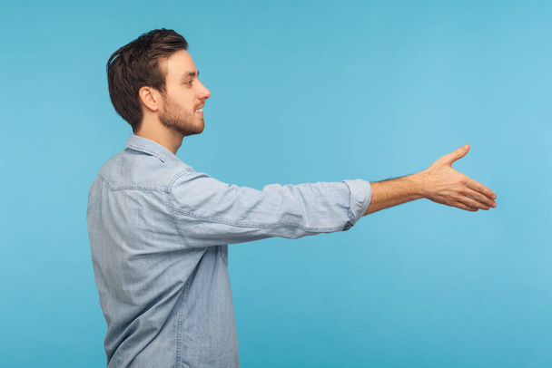 Hi, nice to meet you! Side view of friendly smiling handsome man applicant in worker denim shirt outstretching hand, offering handshake on job interview. indoor studio shot isolated on blue background - Φωτογραφία, εικόνα