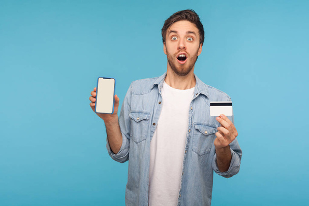 Wow, unbelievable mobile shopping app and online payment! Portrait of amazed man in worker denim shirt showing cell phone and credit card, looking shocked. studio shot isolated on blue background - Photo, image