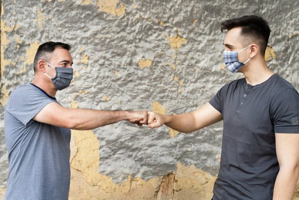 Dwóch mężczyzn w maskach ochronnych witających przed ścianą - Nowy normalny uścisk dłoni uderzenie pięści, aby zapobiec kontakt i rozprzestrzenianiu się wirusa w covid-19 pandemii koronawirusa - Koncepcja odległości społecznej - Zdjęcie, obraz
