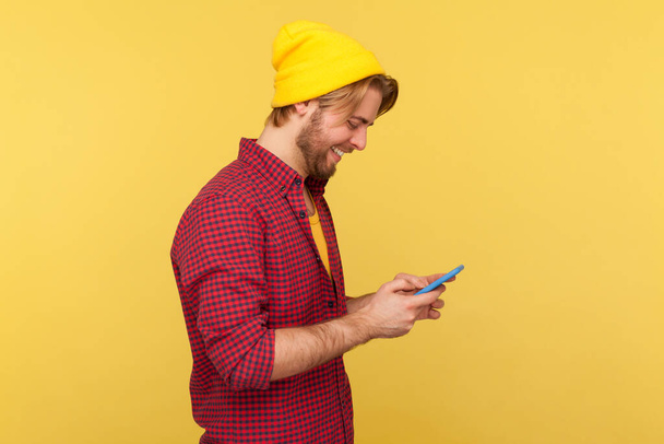 Side view happy trendy guy in hipster beanie hat and shirt using cell phone, texting or reading message, smiling while chatting in social network, enjoying mobile app. indoor studio shot isolated - 写真・画像
