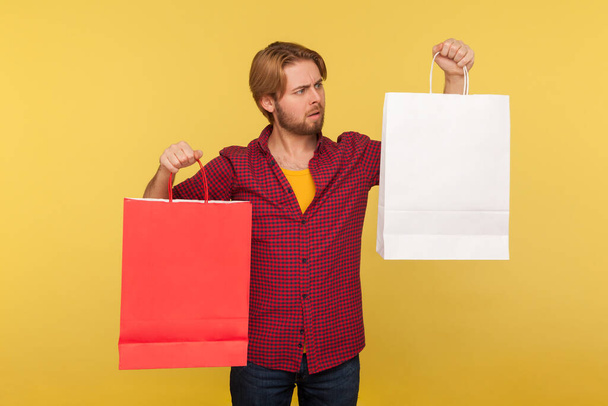 Confused shopper guy in checkered shirt choosing between two shopping bags with mock up empty advertising area, pondering thinking looking at package. indoor studio shot isolated on yellow background - Foto, afbeelding