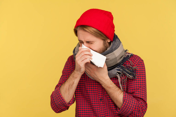Sick guy in hat and warm scarf coughing sneezing napkin, feeling unwell with runny nose, sore throat, suffering allergy or seasonal influenza symptoms. indoor studio shot isolated on yellow background - Foto, afbeelding