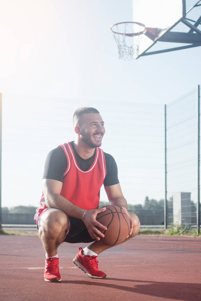 The young smiling basketball player with basketball ball is squatting on playground. - Foto, Bild
