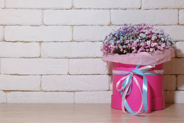 a bouquet of decorative wild flowers in a pink, round, gift box with colorful ribbons stands on a wooden table against a wall of white bricks. - Foto, Imagen